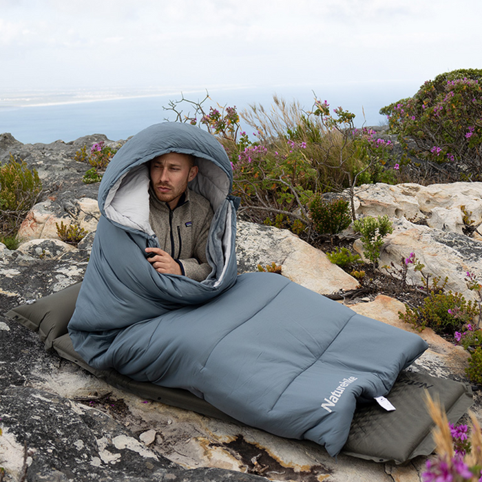 - Vīrietis, sēžot uz klinšainas virsmas, izmantot pelēku Naturehike BE400 USB apsildāmo guļammaisu
- Apkārtne kalnu vidē ar krūmiem un ziediem
- Guļammaisa dizains ar kapuci un zīmolu Naturehike redzams uz guļammaisa
- Guļammaiss nodrošina siltumu un komfortu āra aktivitātēm, piemēram, kempingam un pārgājieniem 

Naturehike