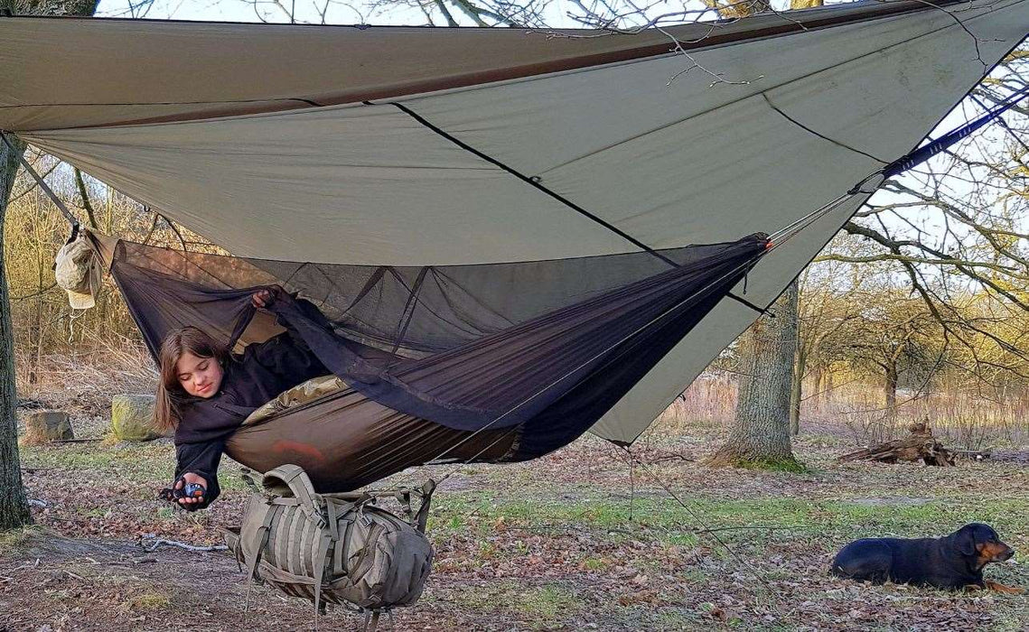 - Persona, kas guļornīcā Bushmen Vagabond Ultralight Hammock ar integrētu moskītu tīklu
- Mājiens, ka guļornīca aizsargā no kukaiņiem
- Koka apkārtne lauva
- Somu un citus piederumus, kas pakārti pie guļornīcas
- Tarp virs guļornīcas papildu aizsardzībai no vides ietekmes
- Bushmen