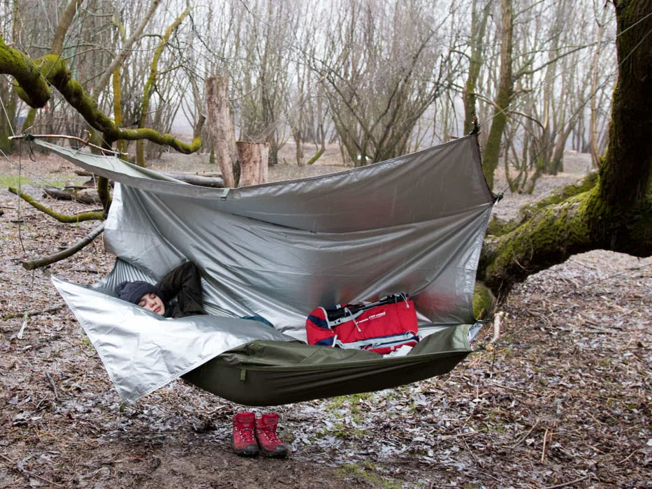 - Stiprs, ūdensizturīgs THERMO Hammock lietus pārsegs ar sildelementu 
- To uzstādīts mežā starp diviem kokiem
- Ar cilvēku, kas guļ ērti guļamtīklā, 
- Sarkans piepūšams matracis un zābaki blakus
- Olīvas krāsas materiāls ar sudraba siltumizolācijas slāni, kas nodrošina papildus siltuma komfortu un UPF 50+ aizsardzību
- Piemērots kempingiem un ārpustelpu izdzīvošanas situācijām
- Bushmen