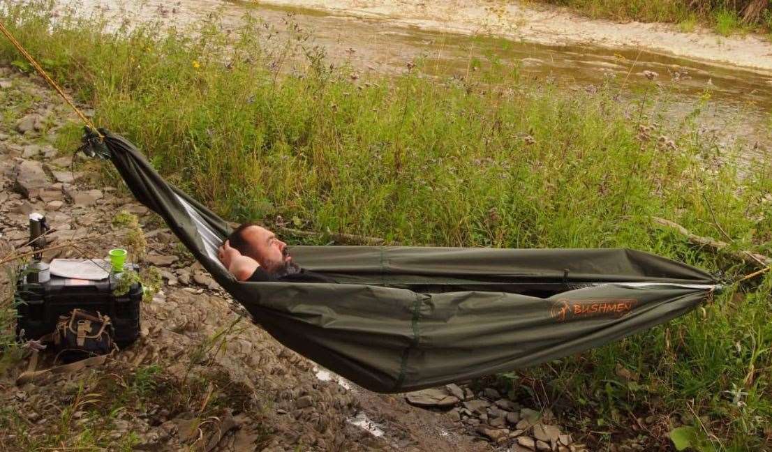 - Cilvēks, kas atpūšas Bushmen THERMO Hammock Tarp 3×3 olīvu krāsā, kas uzstādīta starp kokiem pie upes,
- Bushmen