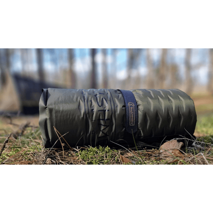 - Bushmen Self Inflating Sleeping Pad FIT 480 iepakotā stāvoklī guļ uz zemes, tumši zaļā krāsā, nostiprināta ar melnu velcro siksnu – Bushmen,
