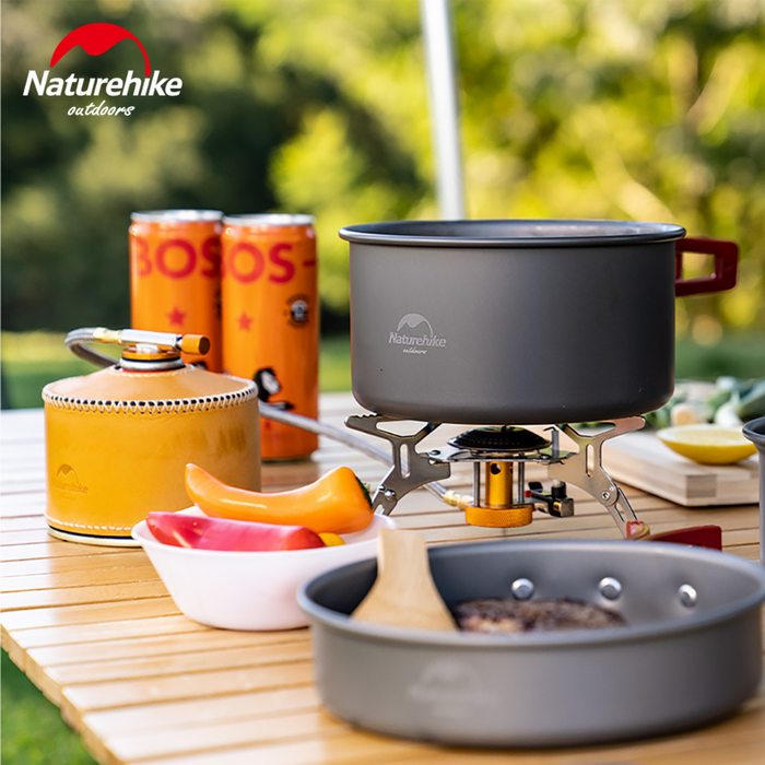 - Naturehike Ultralight Portable Gas Stove novietota uz koka galda ārā, ar sildāmo katlu un pannas blakus
- Piekļauti piederumi un divas dzērienu bundžas novietotas uz galda blakus krāsniņai
- Fona zaļumi norāda uz šīs gāzes plīts piemērotību āra aktivitātēm
- Kompakts dizains un viegli pārnēsājams risinājums ēdiena gatavošanai kempingā vai piknikā
- Zīmola logotips redzams uz produkta katla
- Naturehike