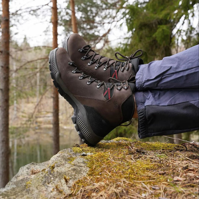 Pārgājienu zābaki Alpina Prima Mid Classic brūns nubuks uz kājām cilvēkam, kurš sež un tur vienu zābaku uz otra uz akmeņa